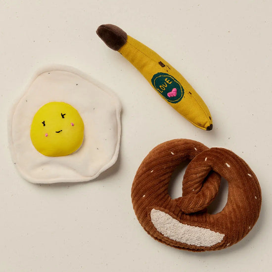 Three plush toys shaped like an egg, banana, and pretzel on a light background