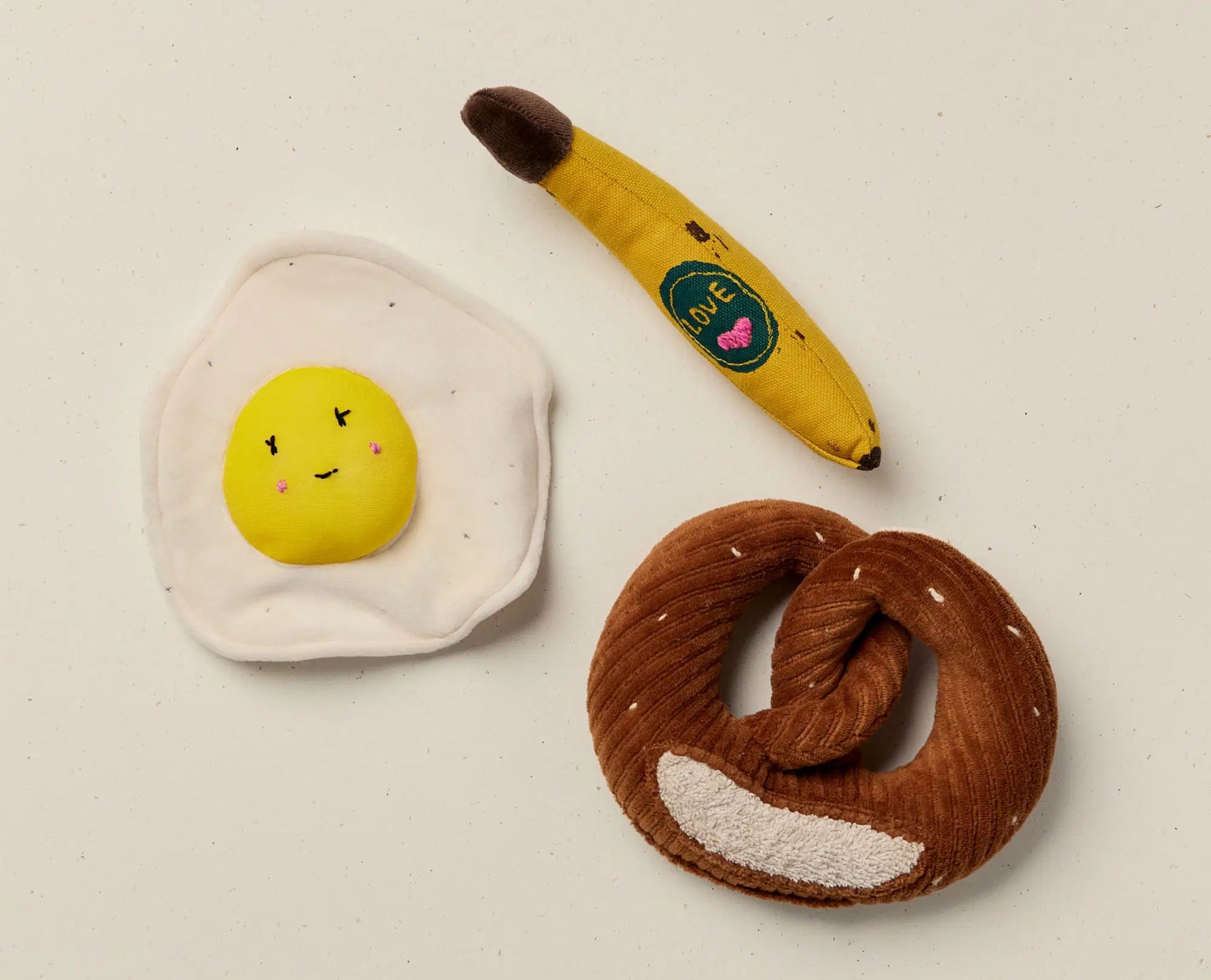 Three plush toys shaped like an egg, banana, and pretzel on a light background