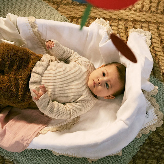 Baby lying in a crib with toys, wearing a beige sweater.