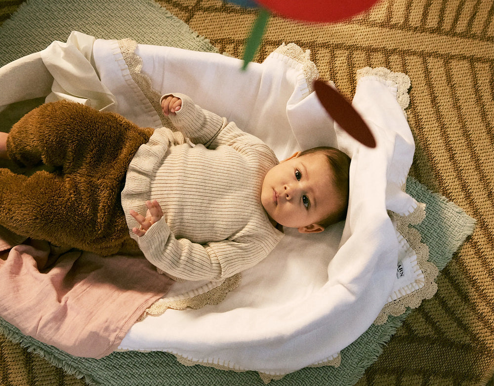 Baby lying in a crib with toys, wearing a beige sweater.