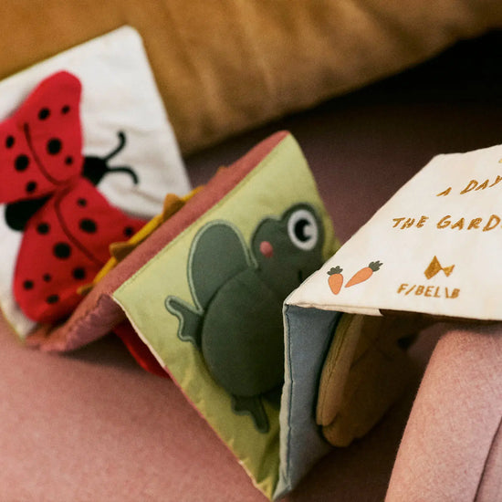 Children's fabric books with a ladybug, frog, and garden-themed pages on a pink surface.
