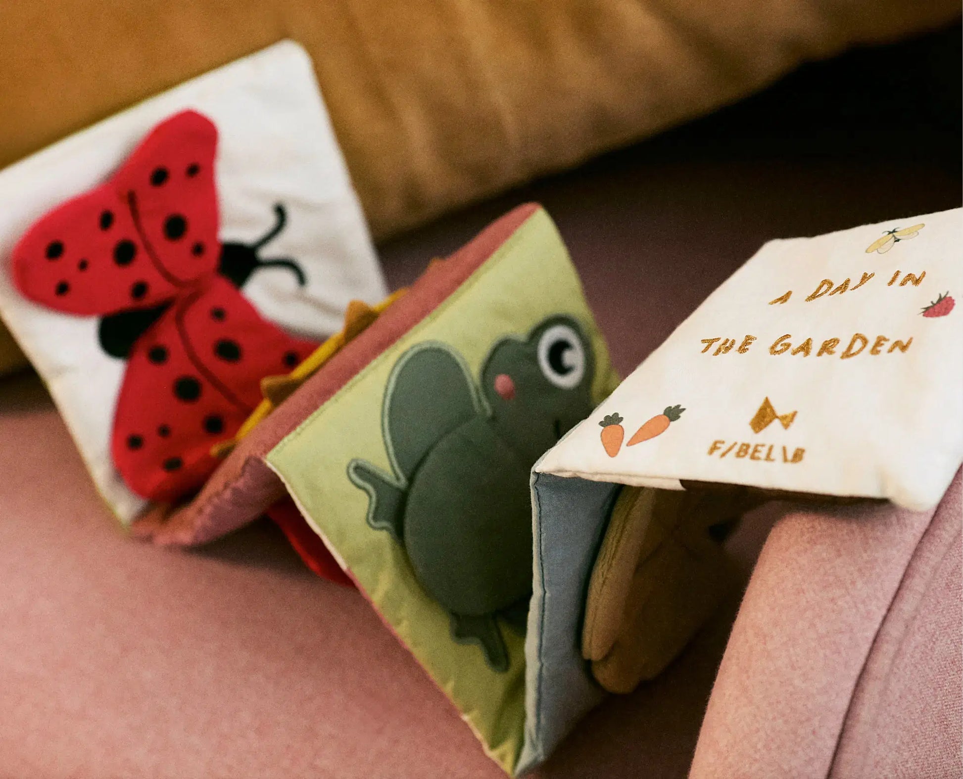 Children's fabric books with a ladybug, frog, and garden-themed pages on a pink surface.