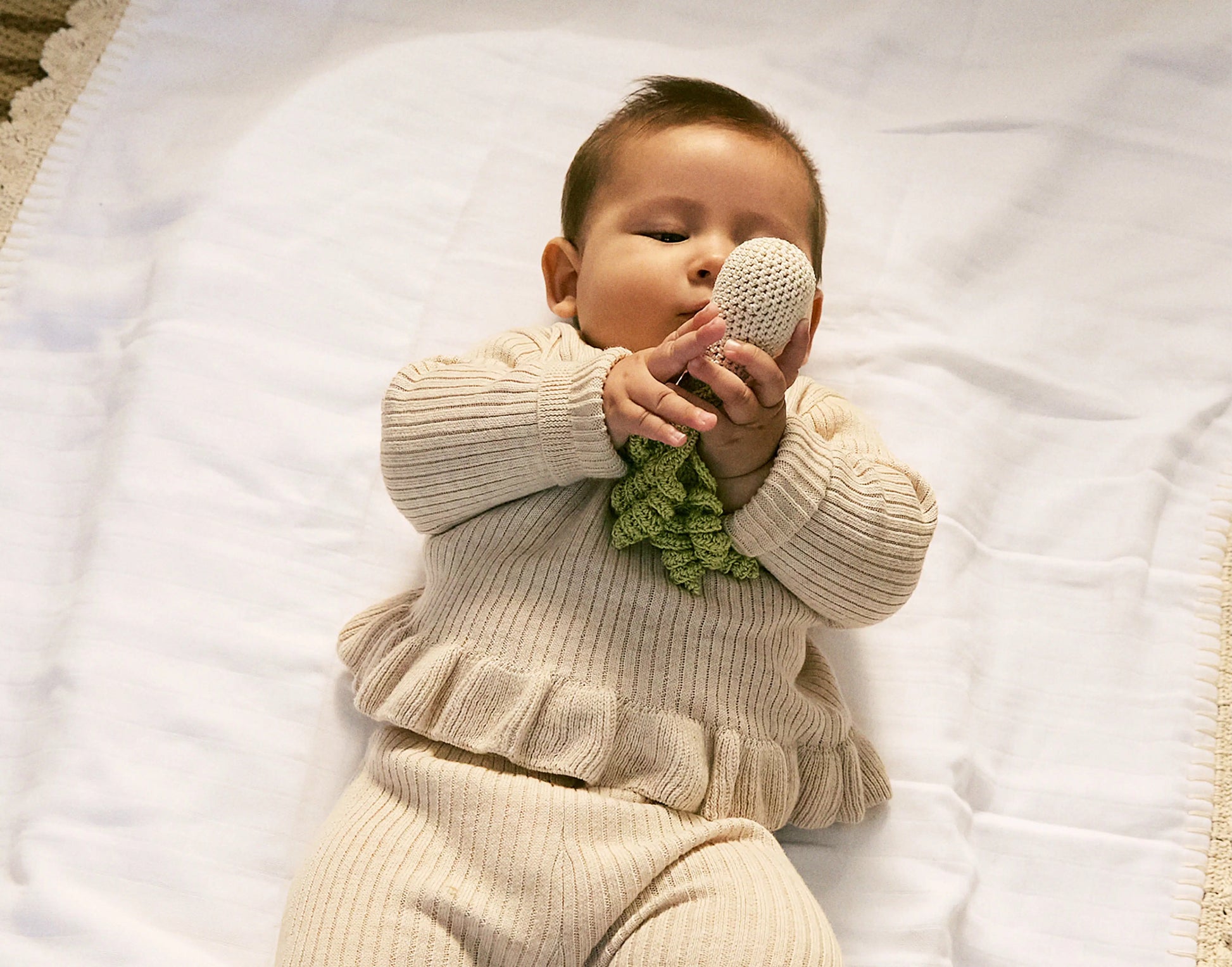 Baby in a beige outfit holding a knitted toy on a white blanket
