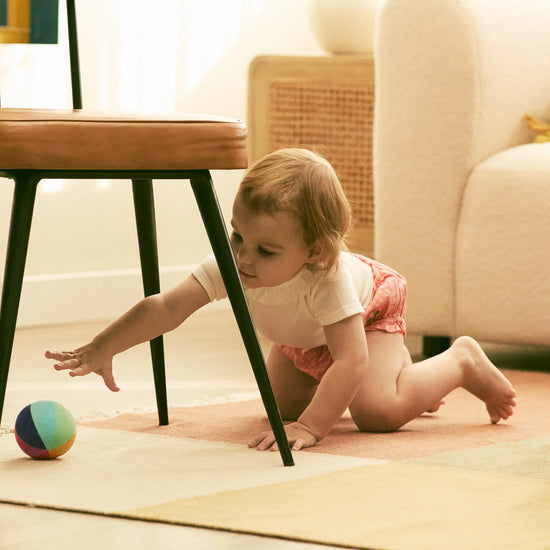 Baby crawling on a rug in a living room with a chair and colorful ball.