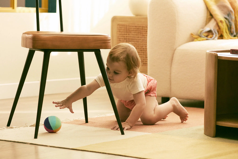 Baby crawling on a rug in a living room with a chair and colorful ball.